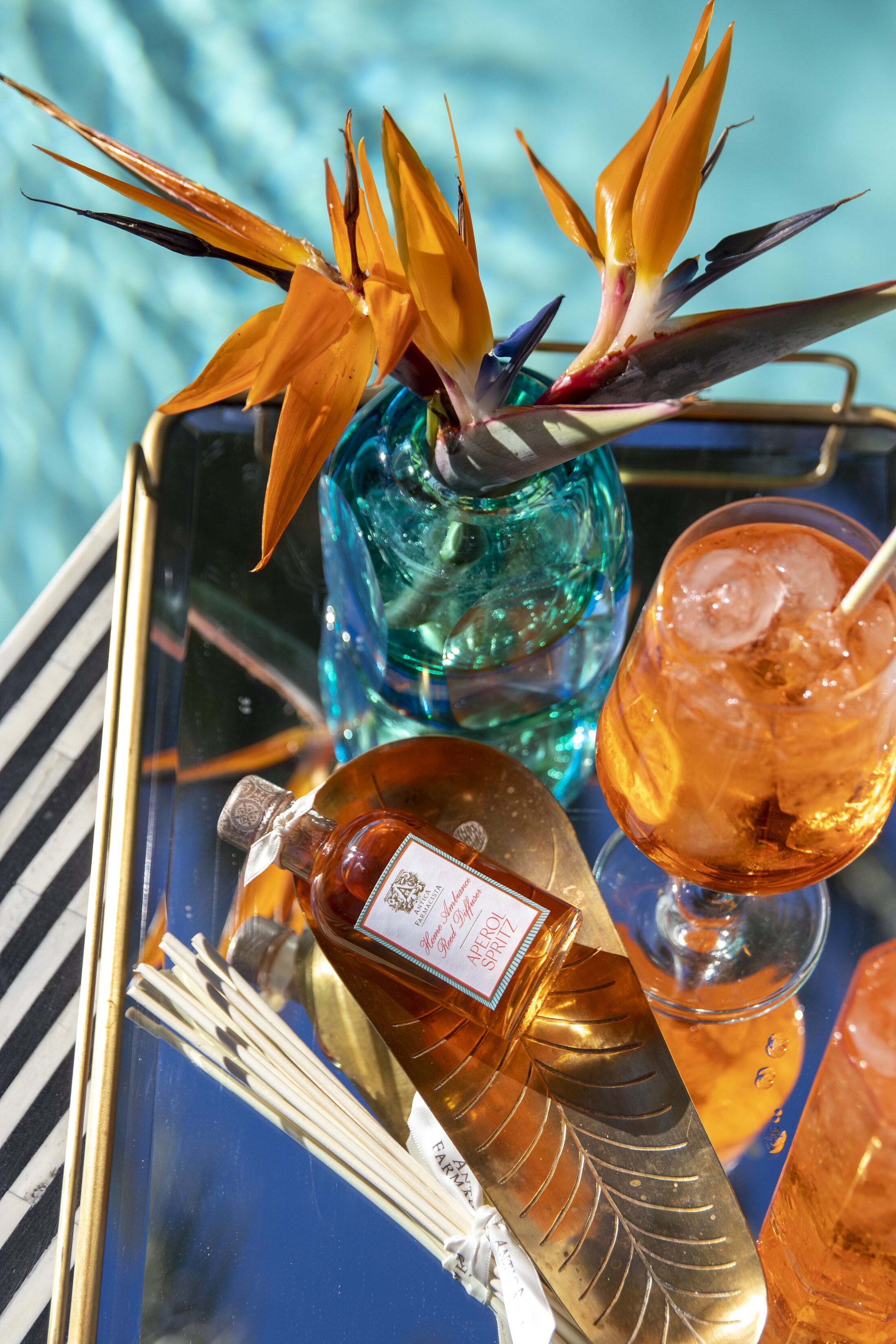 An overhead mirrored tray with birds of paradise in a turquoise vase, the gold-wrapped Aperol Spritz fine fragrance, and spritzes in stemmed glasses.