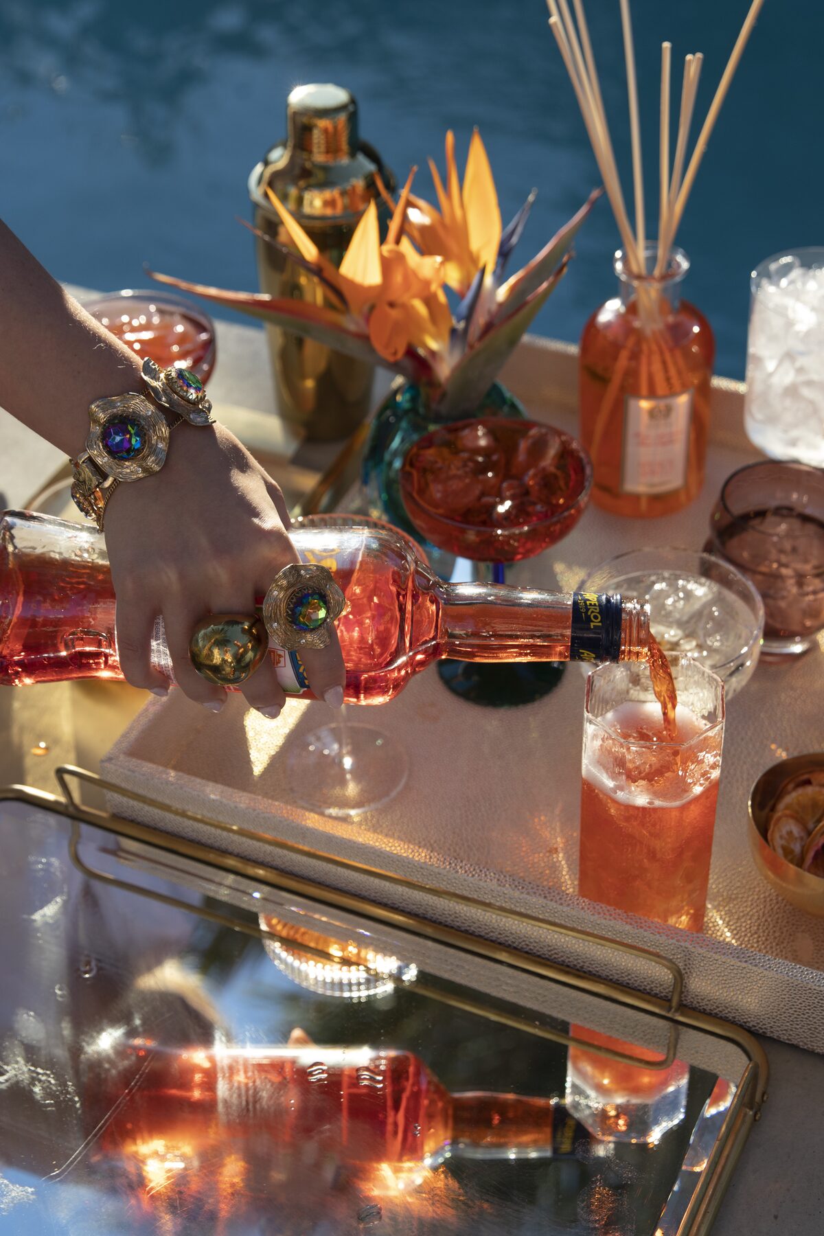 Aperol mid-pour into an iced glass at the poolside bar cart, ringed hands and birds of paradise behind.