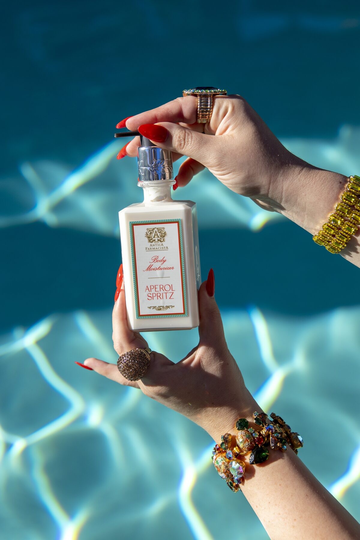 The body moisturiser held in both hands over the pool, nails painted Aperol red.