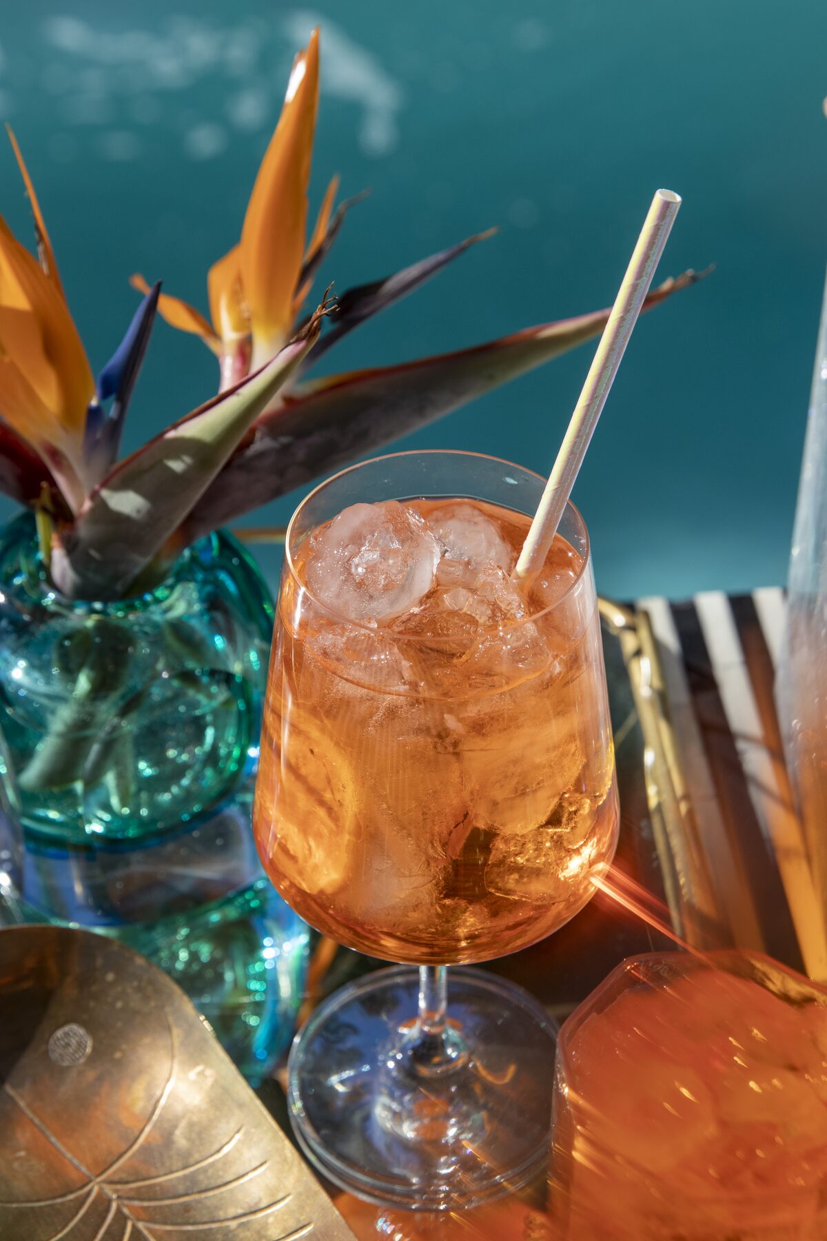 A spritz in a stemmed glass beneath birds of paradise in a teal vase, straw catching the sun.