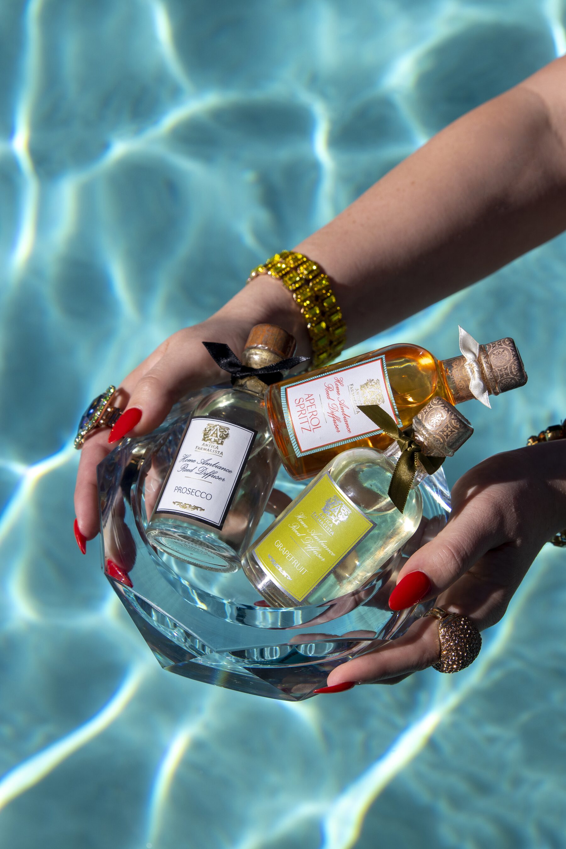 The Prosecco, Aperol Spritz, and Damascena trio held in a faceted crystal bowl over sunlit pool water, hands stacked with jewels.