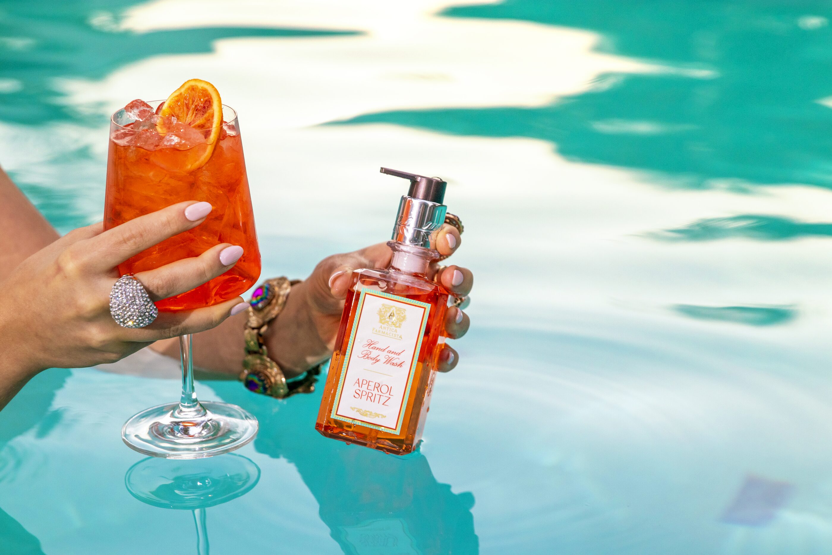 The Aperol Spritz hand wash held at water level in the pool beside a garnished spritz, turquoise water all around.
