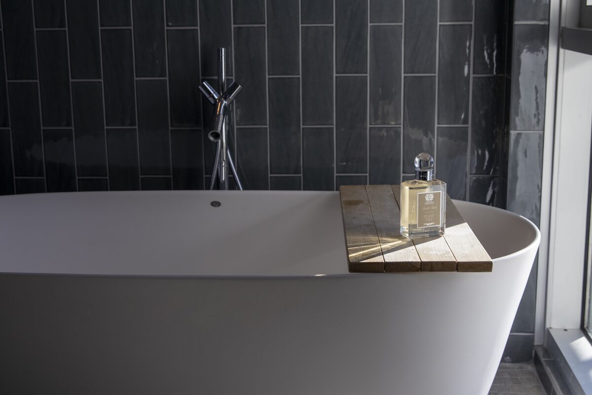 A freestanding tub against dark slate tile, the Desert Sage bath bottle on a teak board across it.