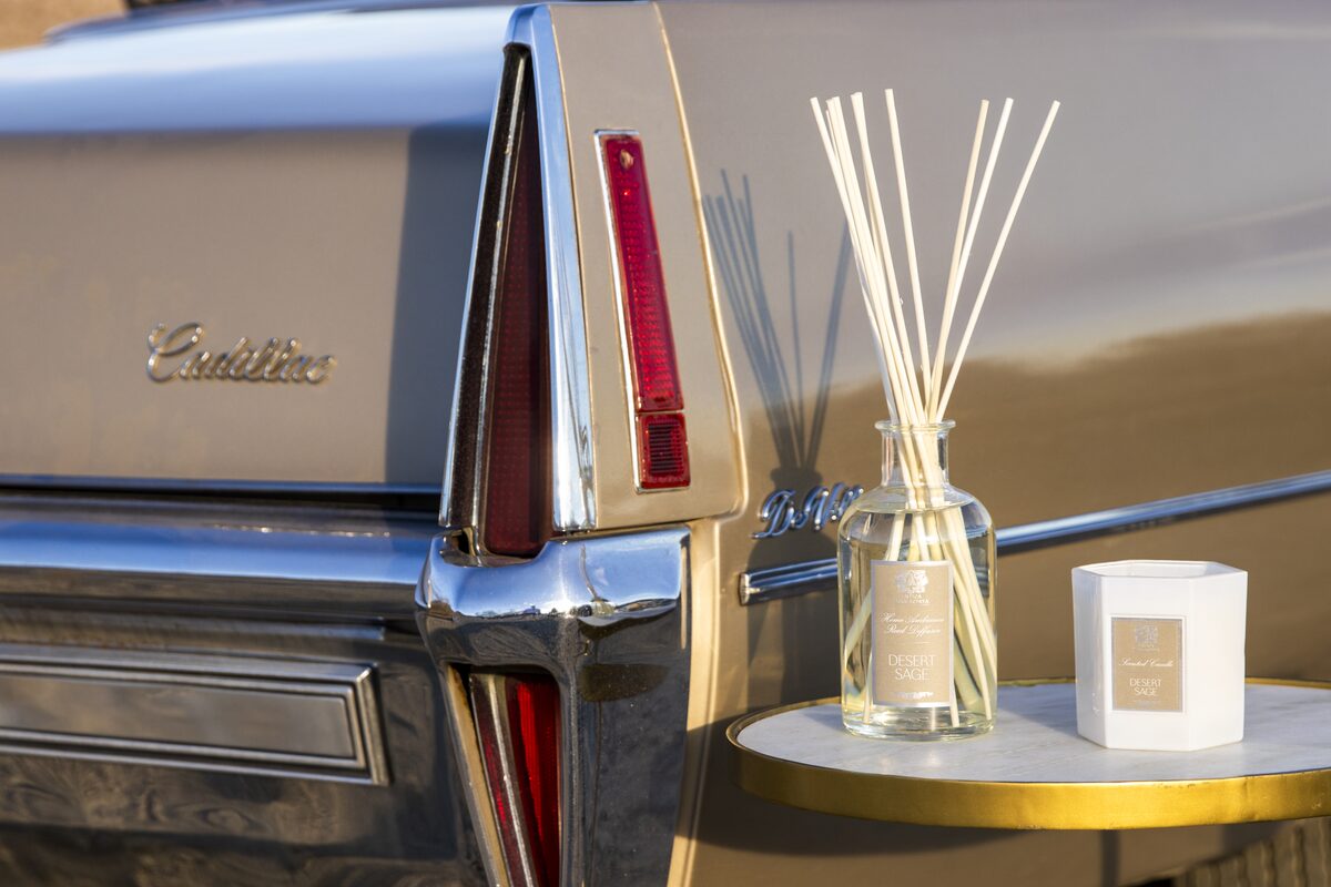 The diffuser and candle on a gold table beside the chrome tail fin of a vintage Cadillac in low desert sun.
