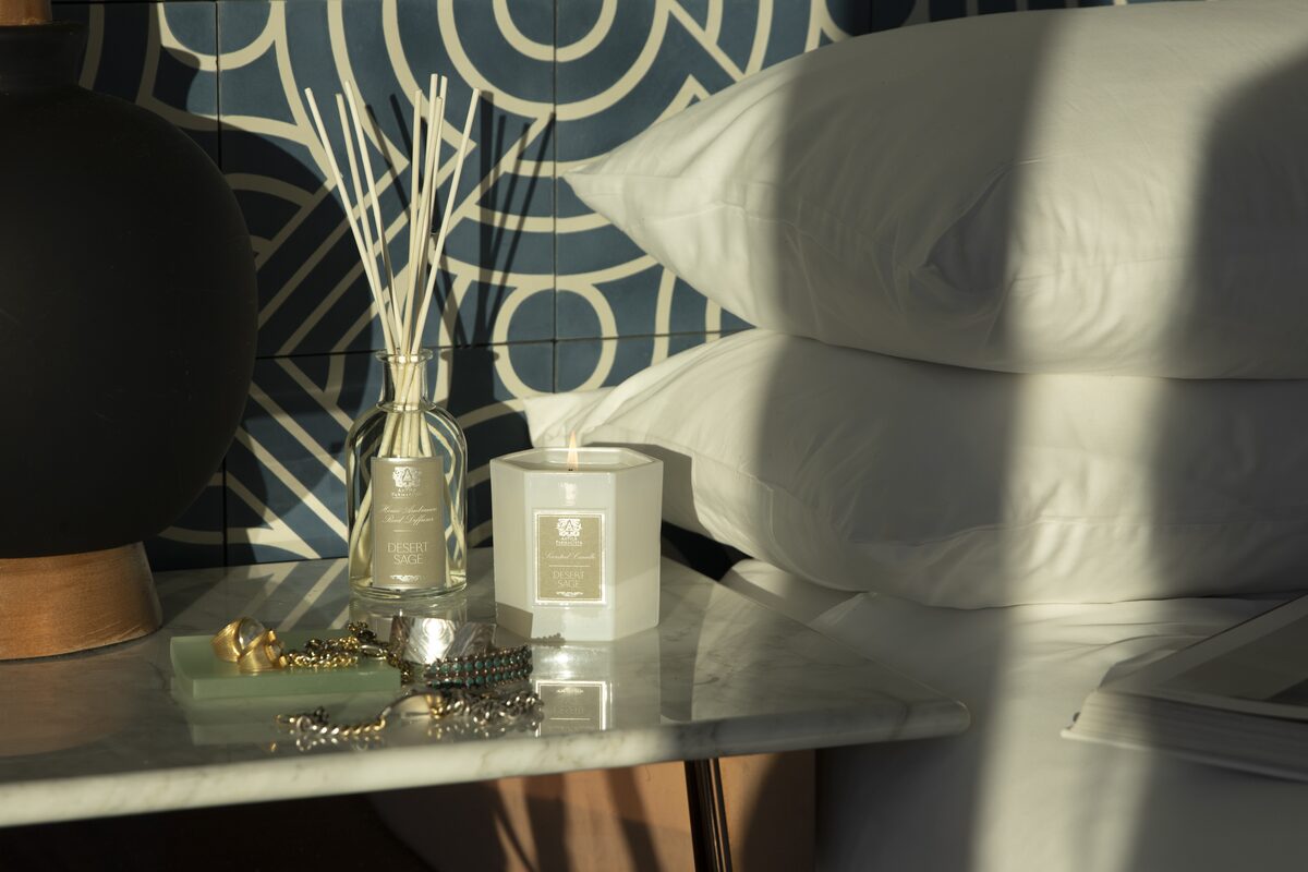 The diffuser and candle on a marble bedside table against a blue patterned wall, stacked white pillows alongside.