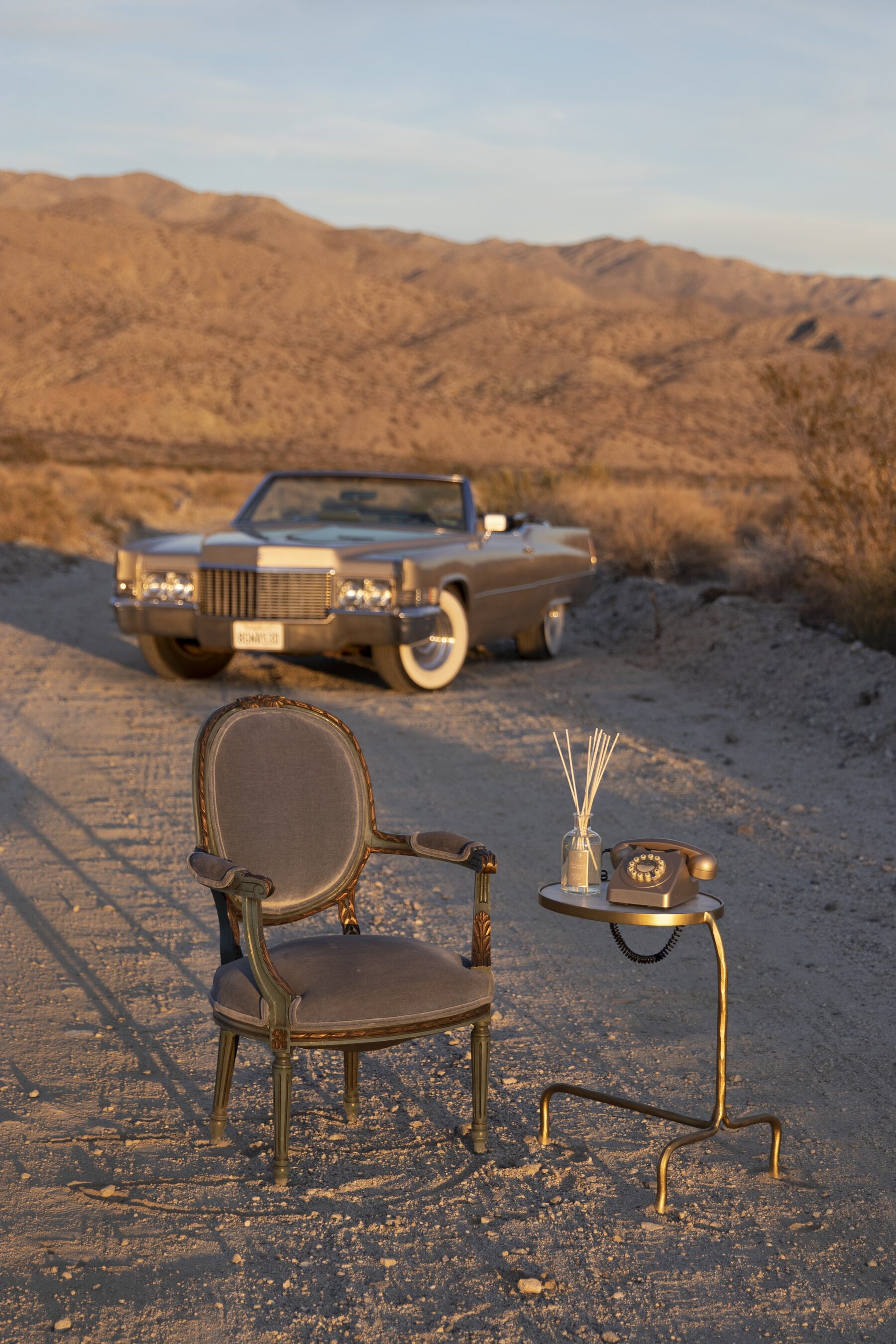 A gilt armchair and gold side table holding a Desert Sage diffuser, set on a desert road with a vintage Cadillac and the mountains beyond at golden hour.