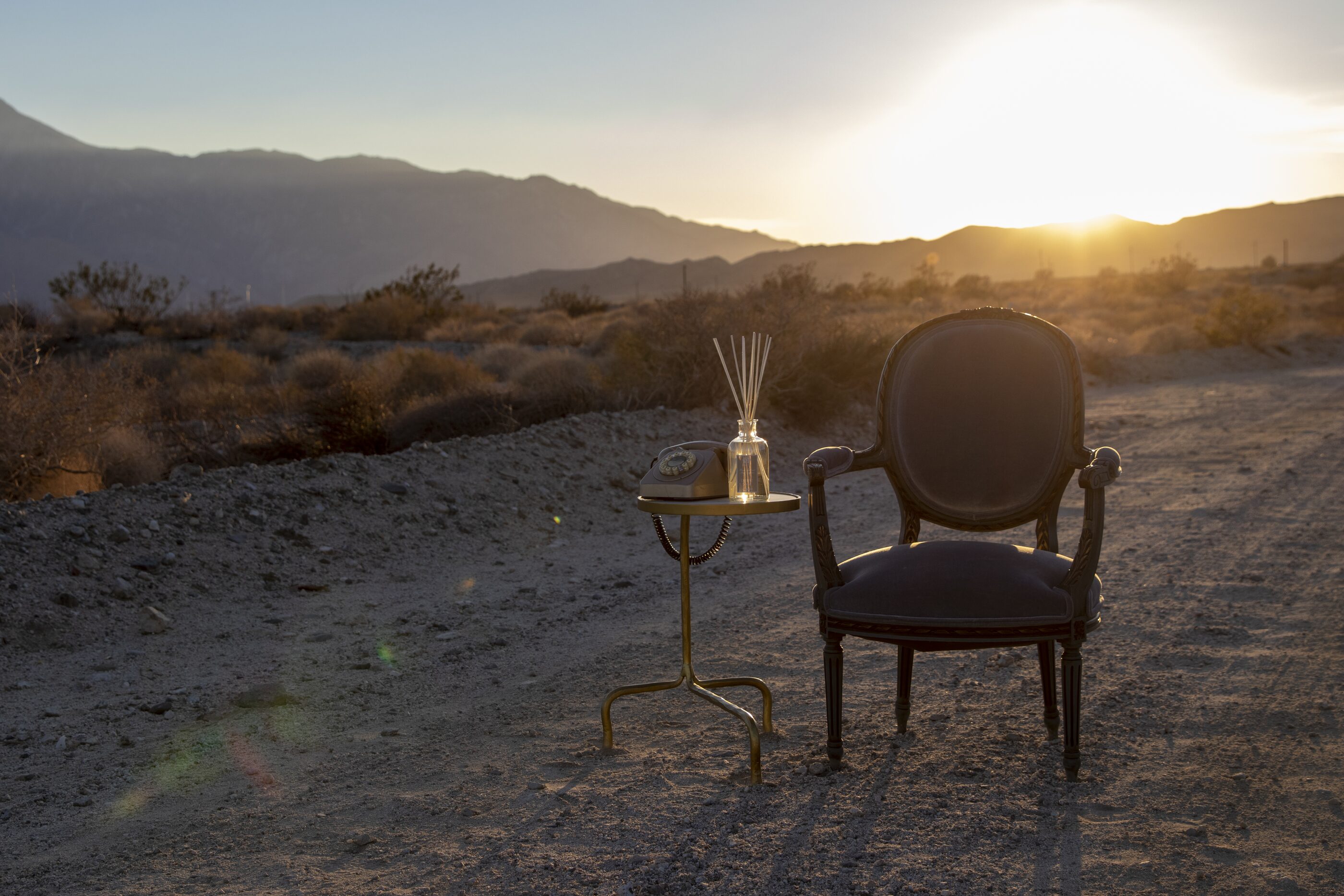 A chair and gold side table set alone on an open desert road as the sun drops behind the mountains.