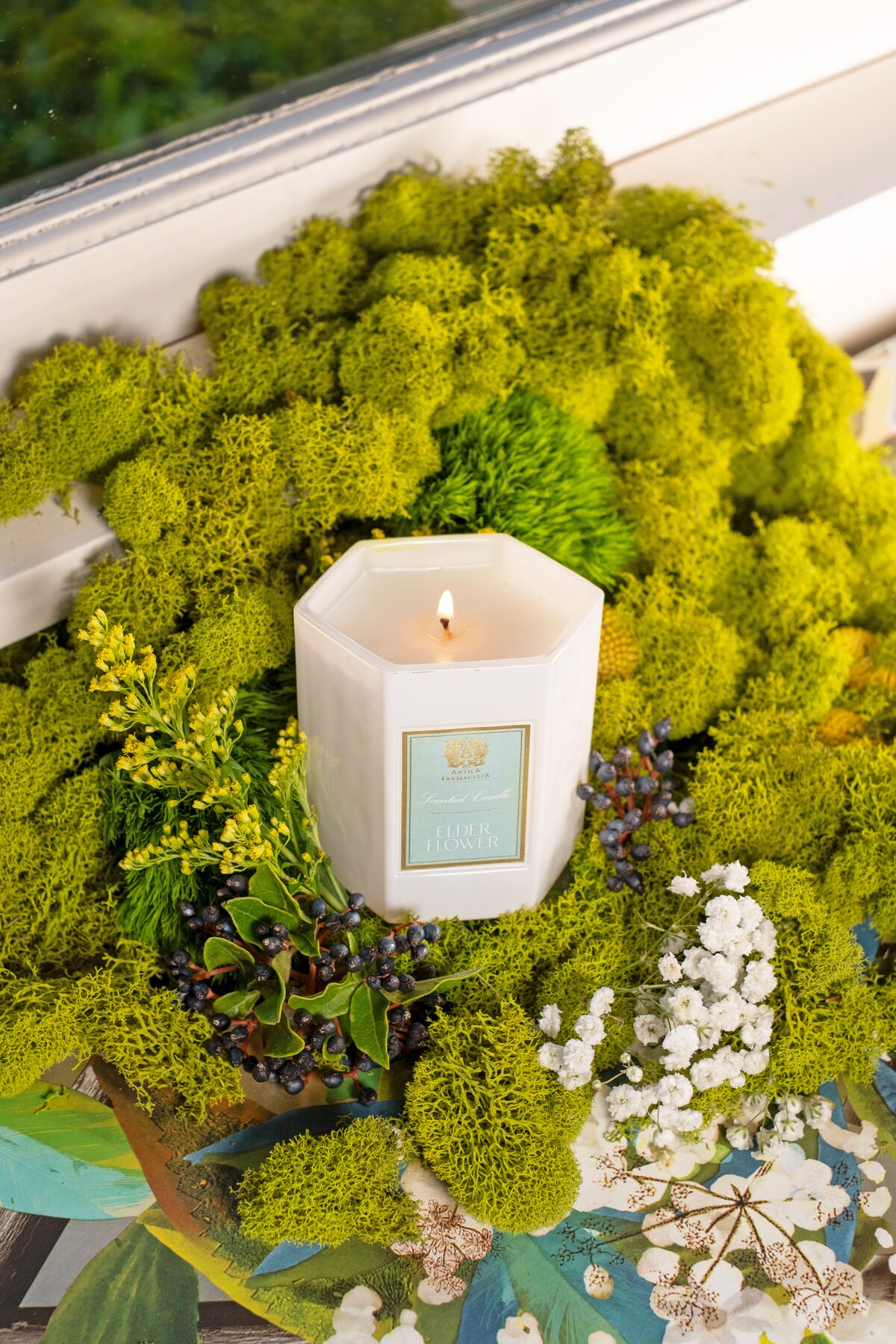 The Elderflower candle lit in a bed of moss and gypsophila at the glasshouse window.