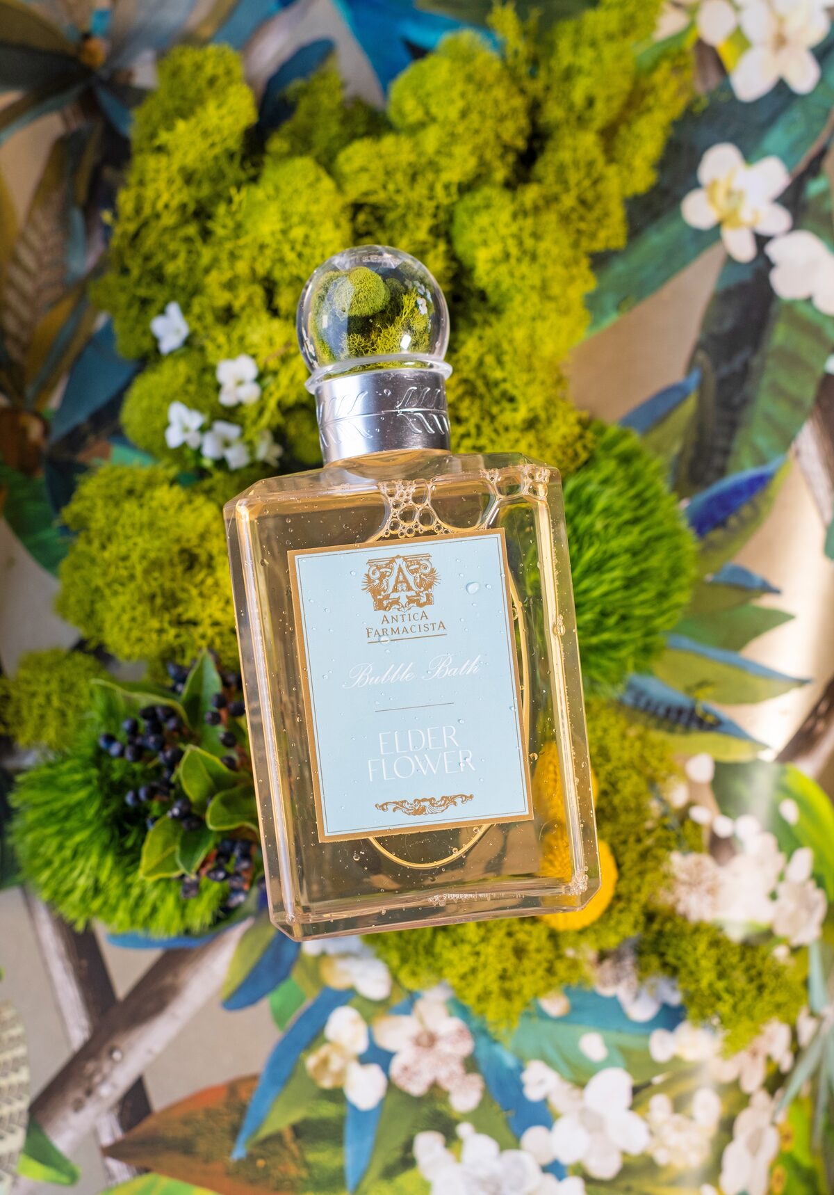 The bubble bath with its crystal-ball cap catching moss in the reflection, collage florals behind.