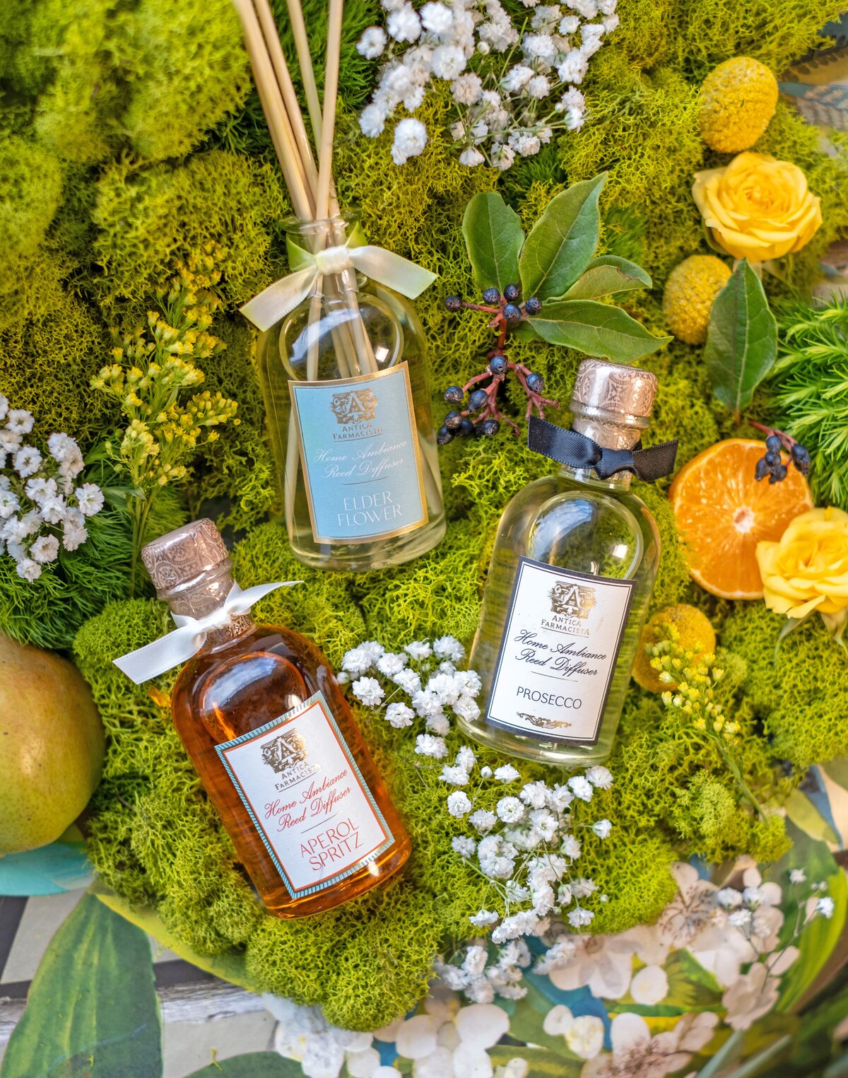 The Elderflower, Aperol Spritz, and Prosecco diffusers nested together in moss with yellow roses and oranges.