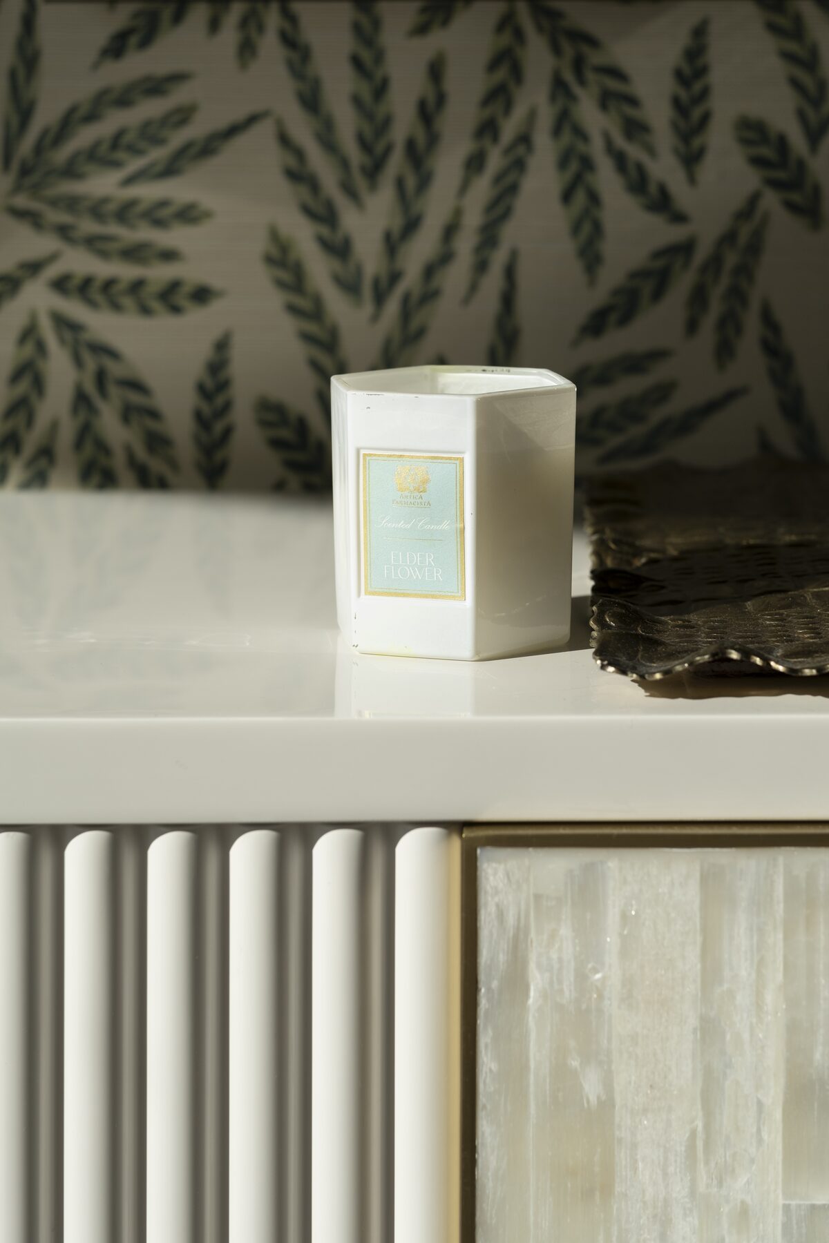 The candle on a white lacquer credenza against fern wallpaper, a bronze leaf dish beside it.