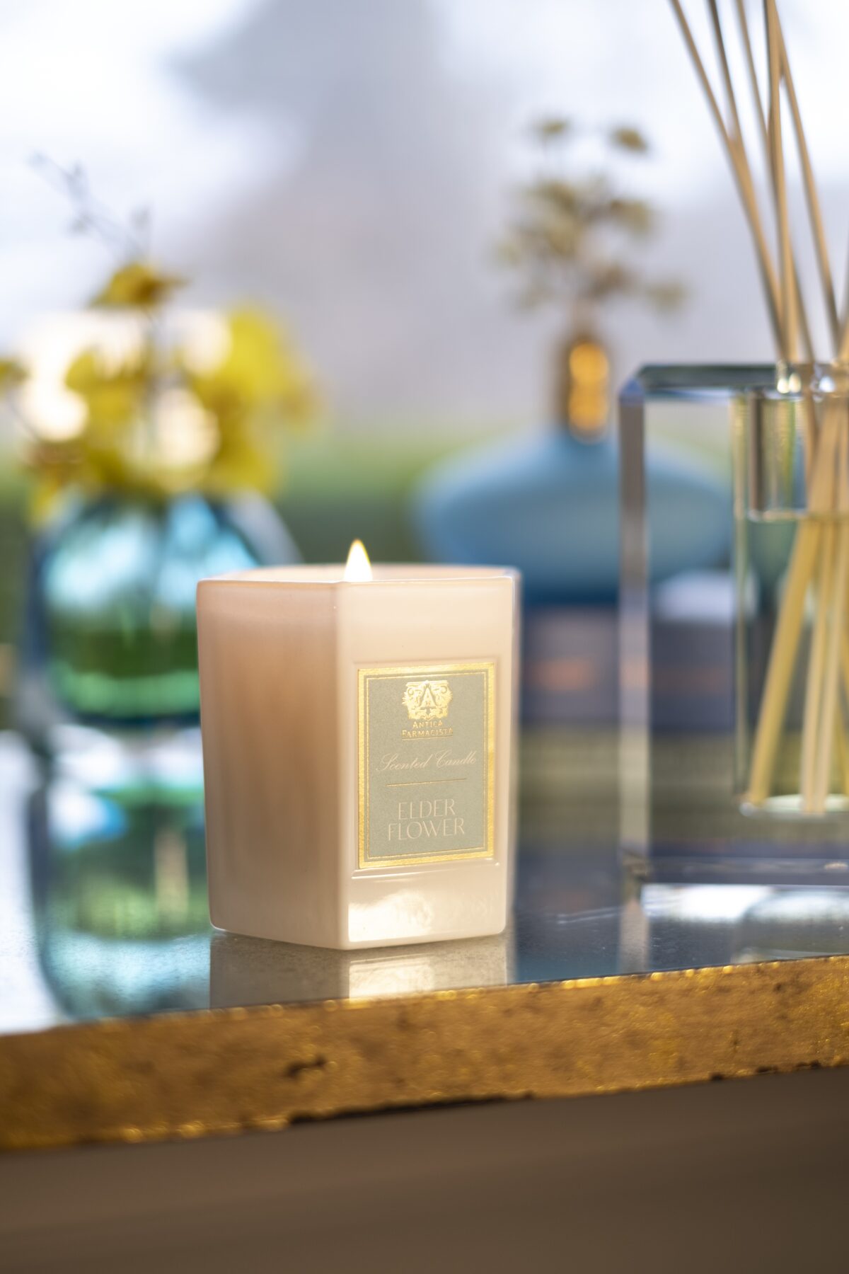 The candle lit on a gold-edged mirror table, blue glass vessels softening behind.