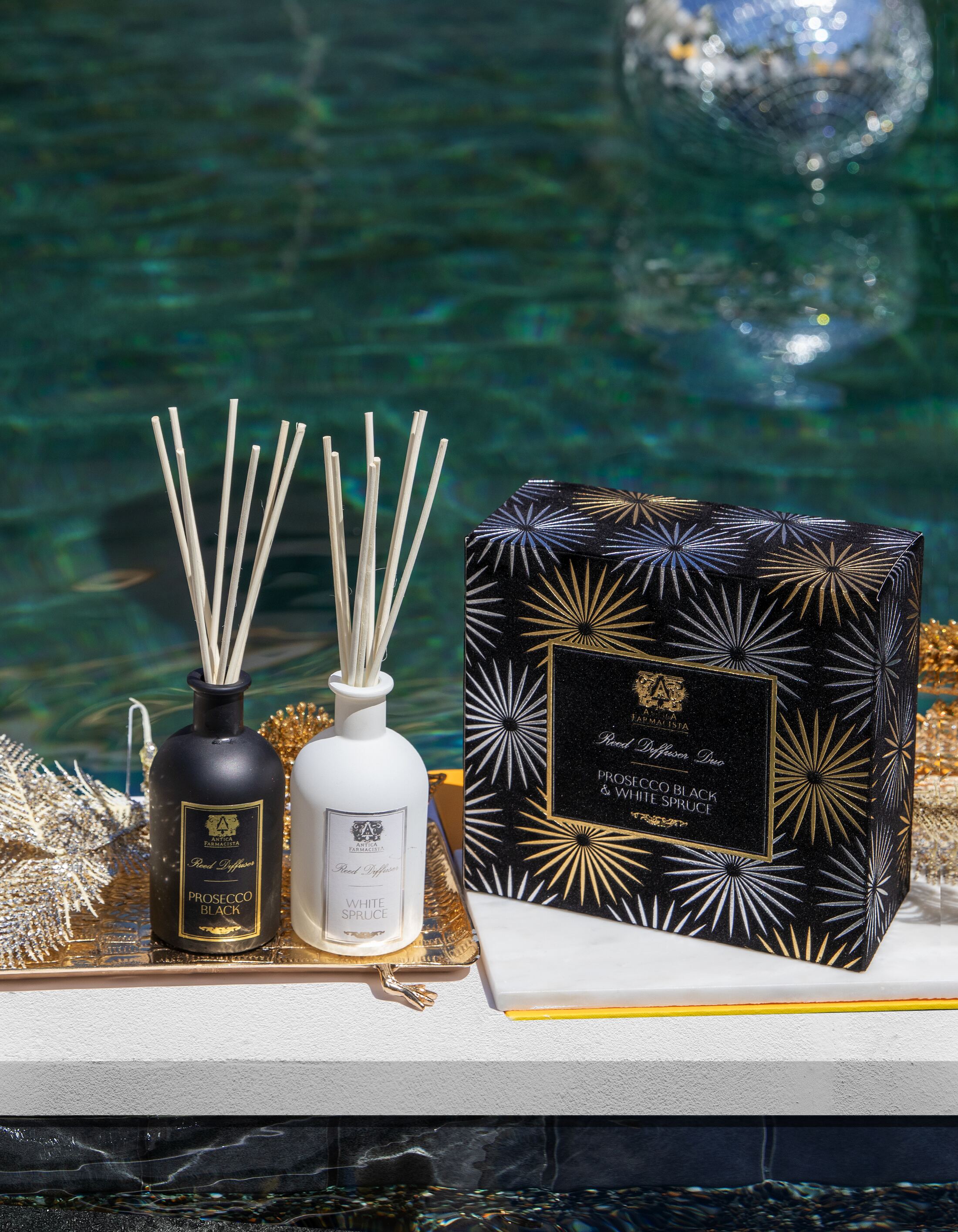 The Prosecco Black and White Spruce diffuser duo on a gold tray at the pool edge, the foiled starburst gift box behind them.