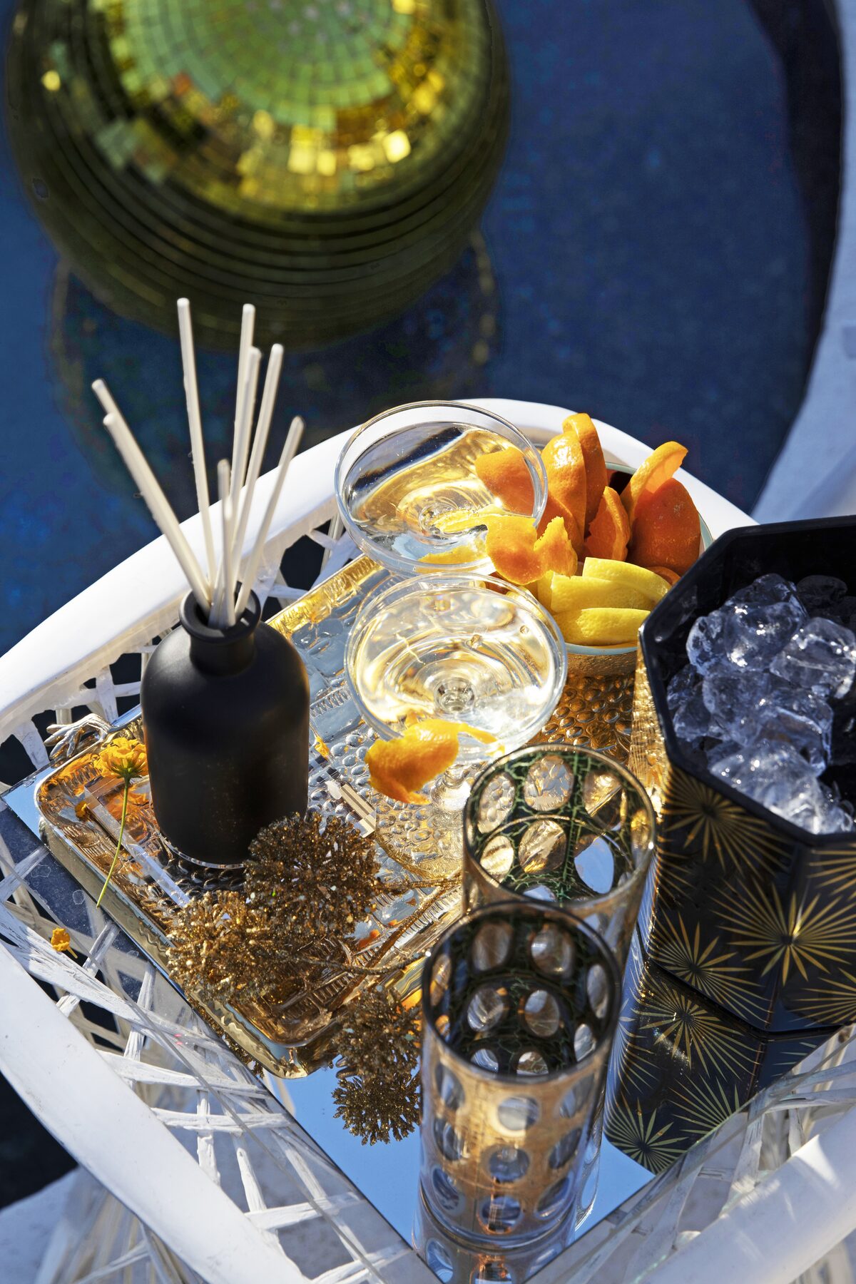 An overhead look at a mirrored cocktail tray by the pool — diffuser, citrus, ice bucket, gold tinsel.