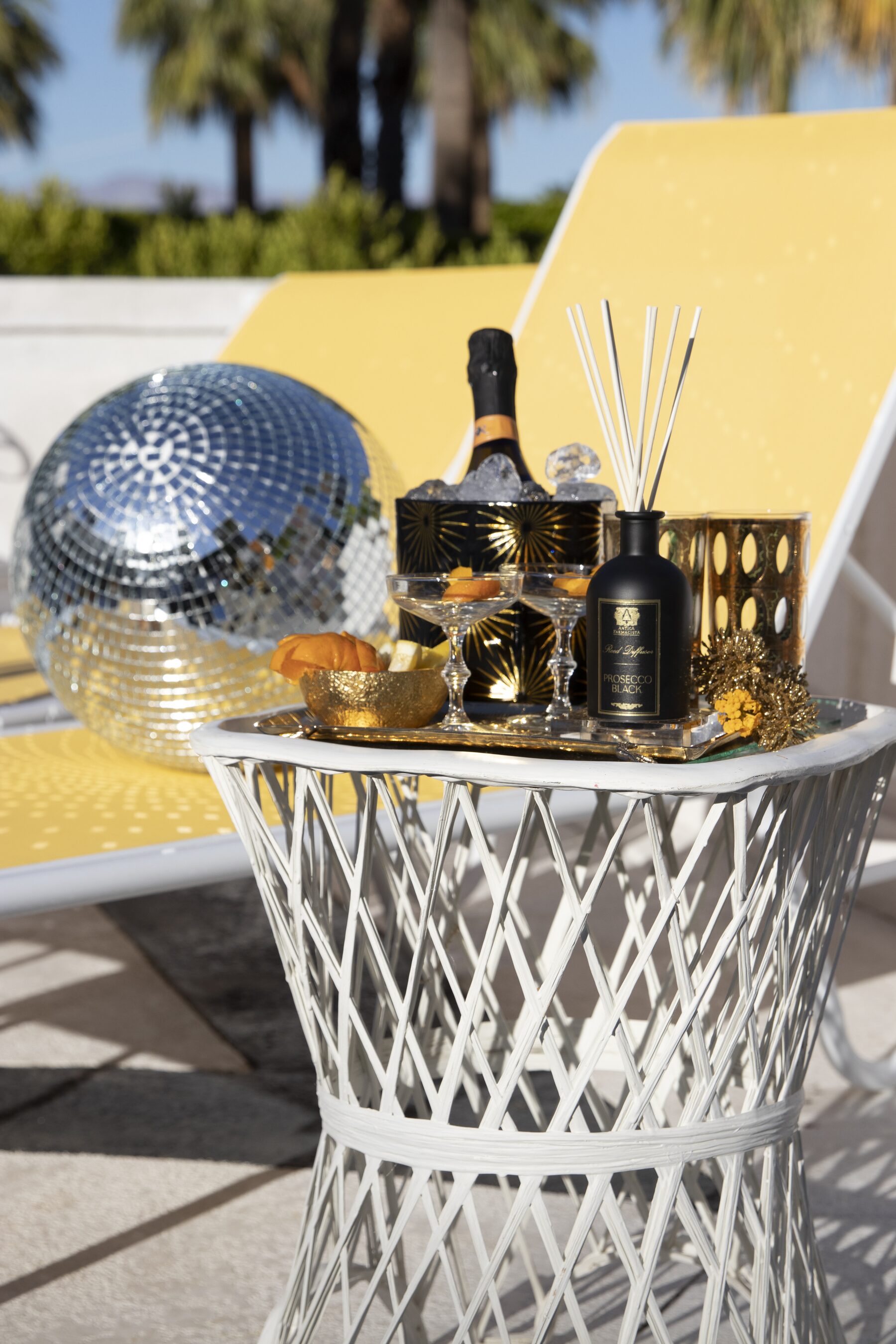 A silver disco ball on a yellow chaise beside a white wicker table of champagne, cocktails, and the Prosecco Black diffuser, palms behind.