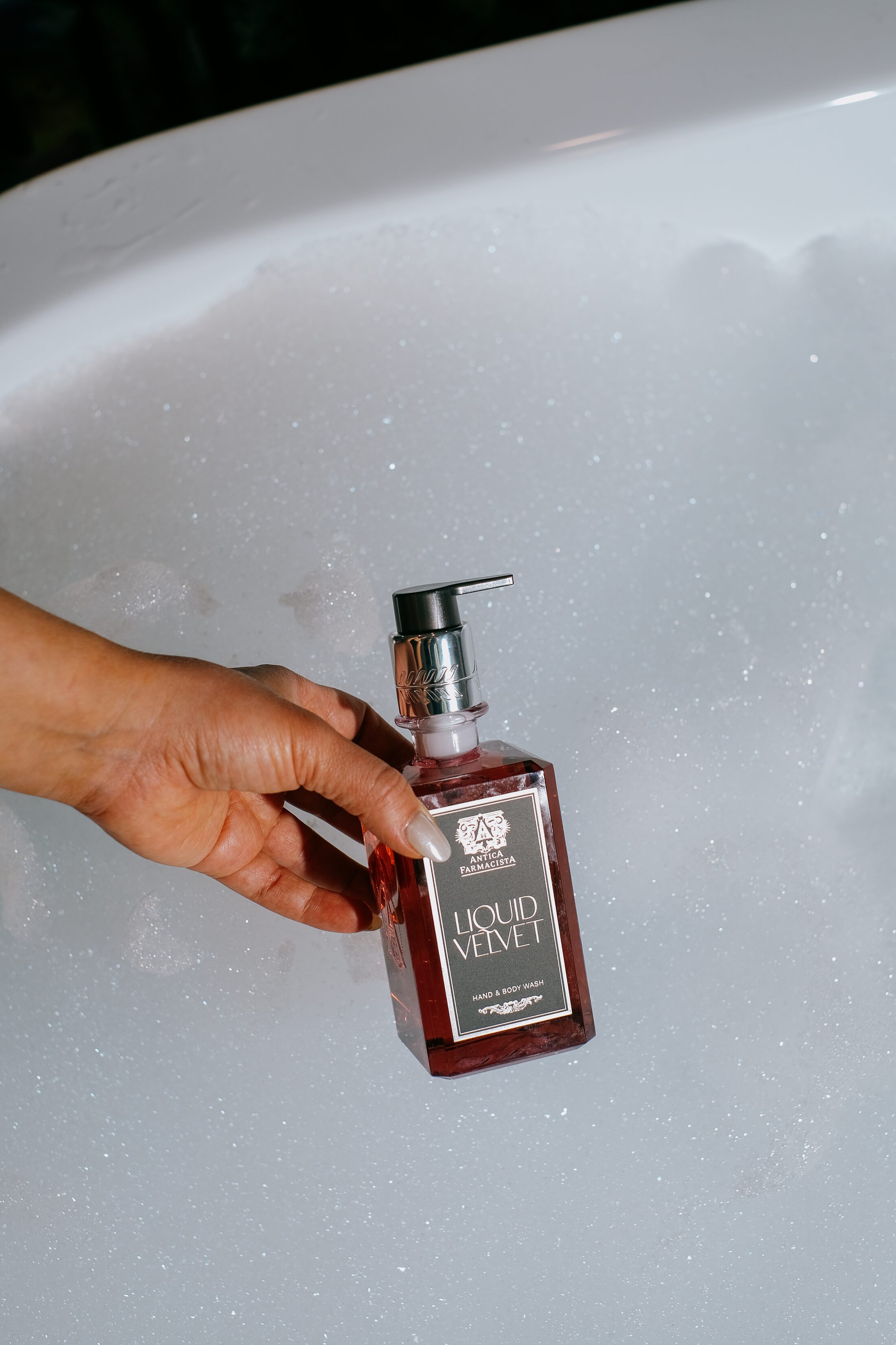 The Liquid Velvet hand and body wash held at the surface of a white bath, bubbles rising.
