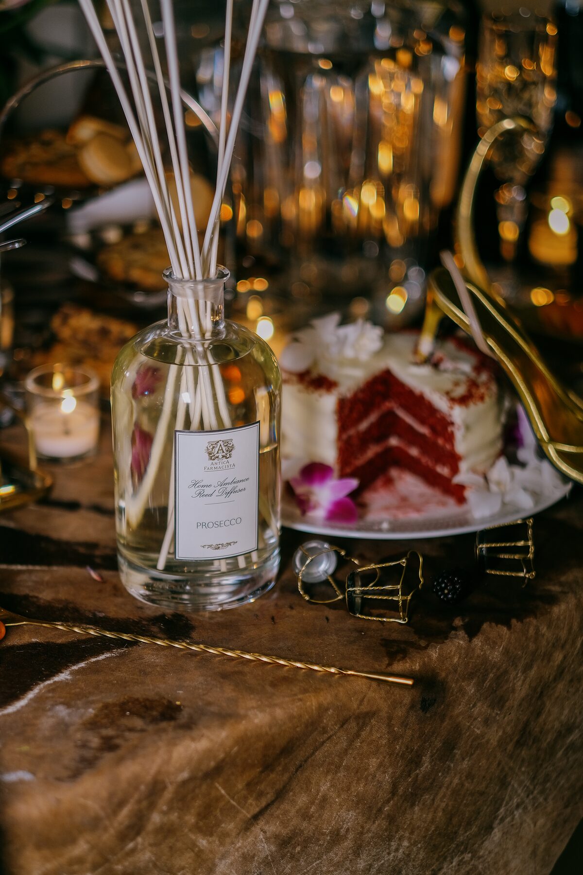 The Prosecco diffuser beside a cut slice of red velvet cake on the fur-draped table, candlelight behind.