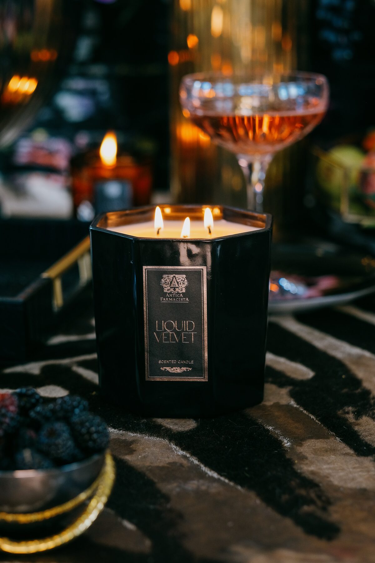 The Liquid Velvet three-wick lit beside a coupe and a bowl of blackberries on zebra hide.