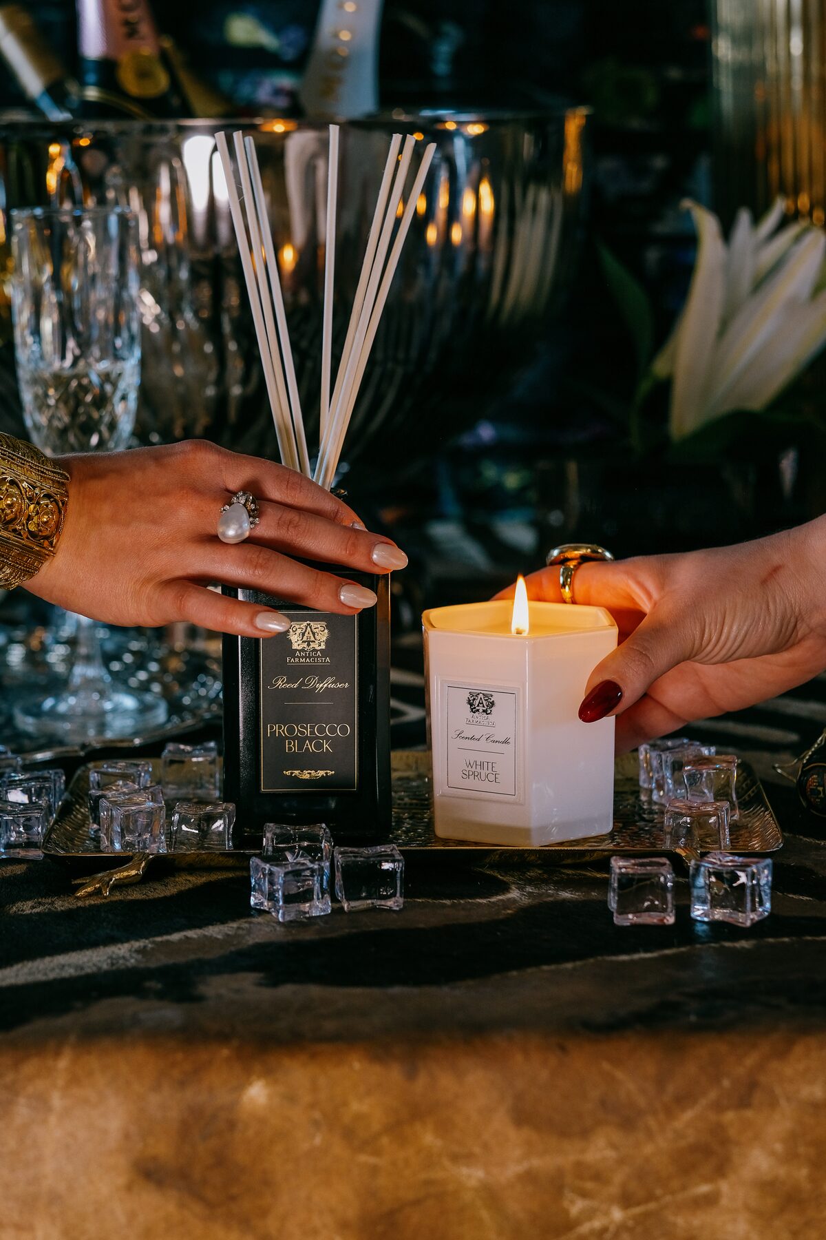 Two hands meet over ice cubes on marble — the Prosecco Black diffuser and a lit White Spruce candle between them.