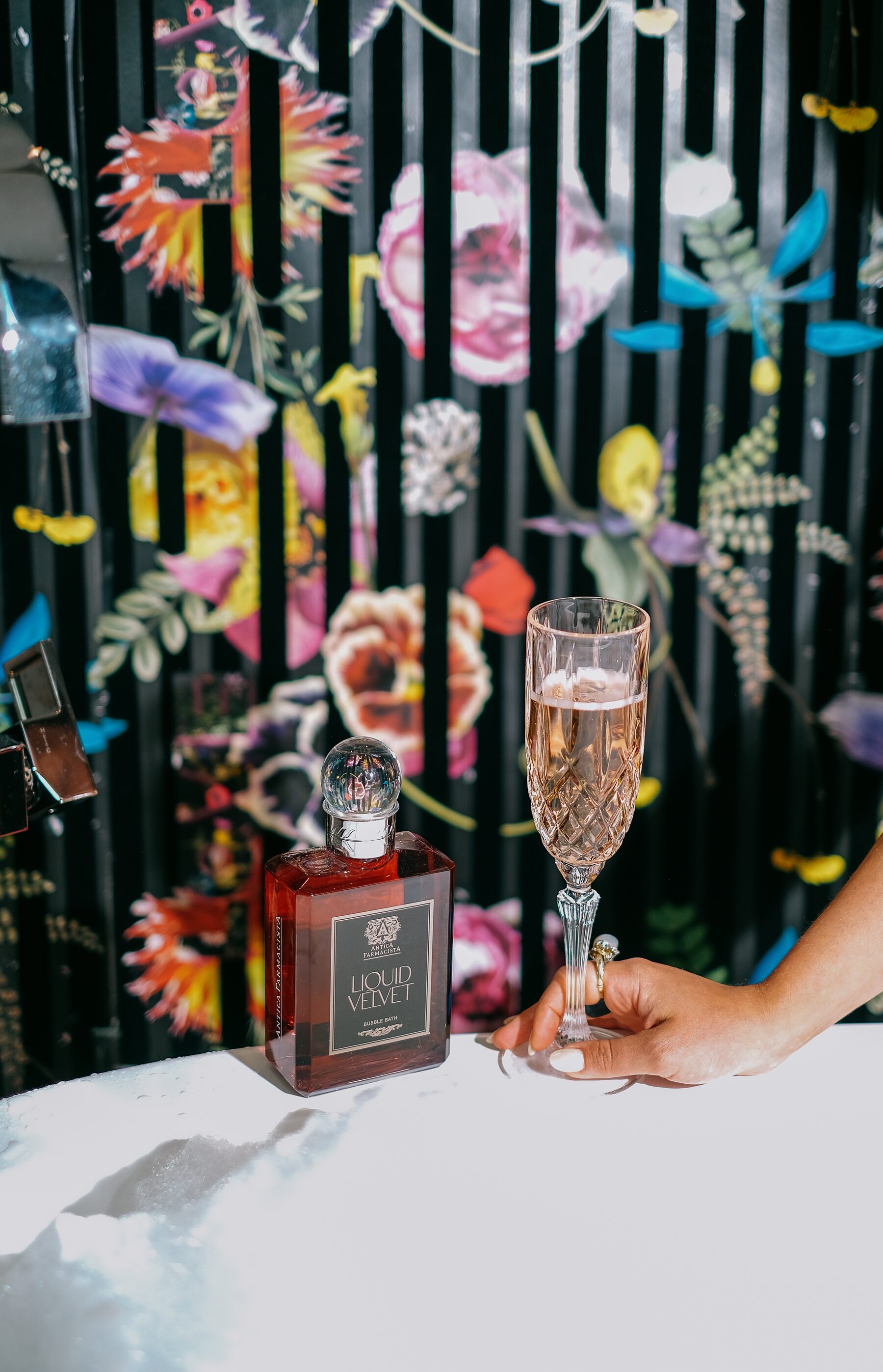The Liquid Velvet bubble bath in its garnet bottle against psychedelic florals, a champagne flute catching the light beside it.
