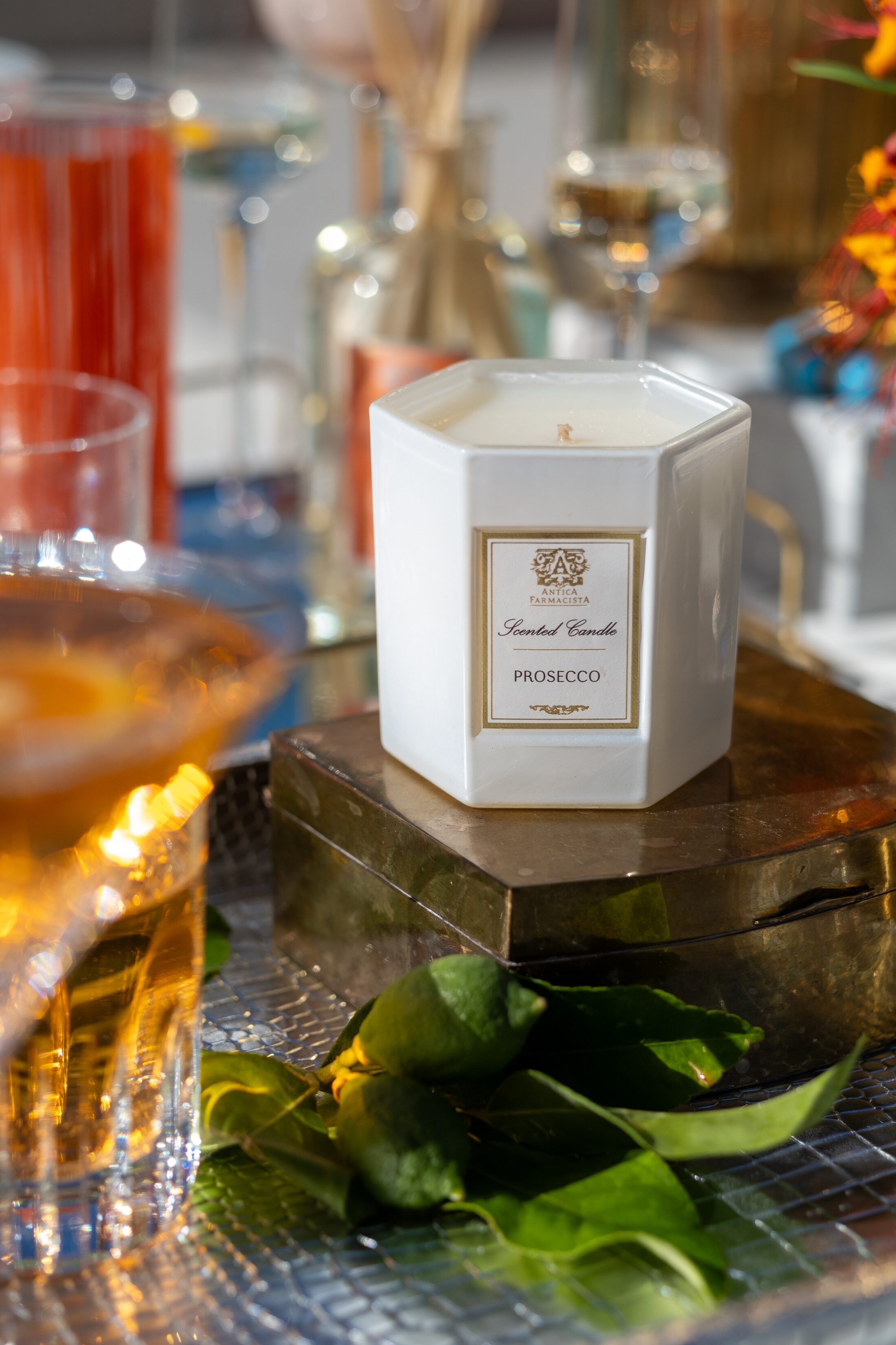 The Prosecco candle on a gold box beside an aperitivo in cut glass, green leaves and pool light behind.