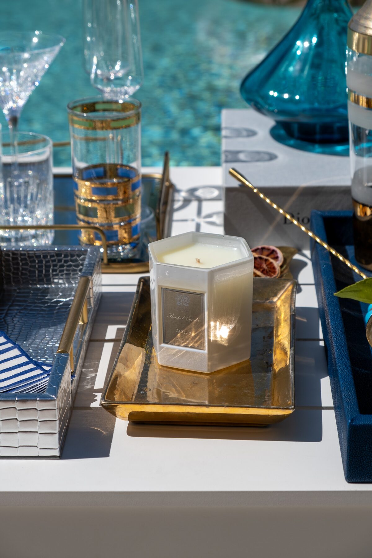 A candle flickering on a silver tray in full daylight, teal glass beyond.