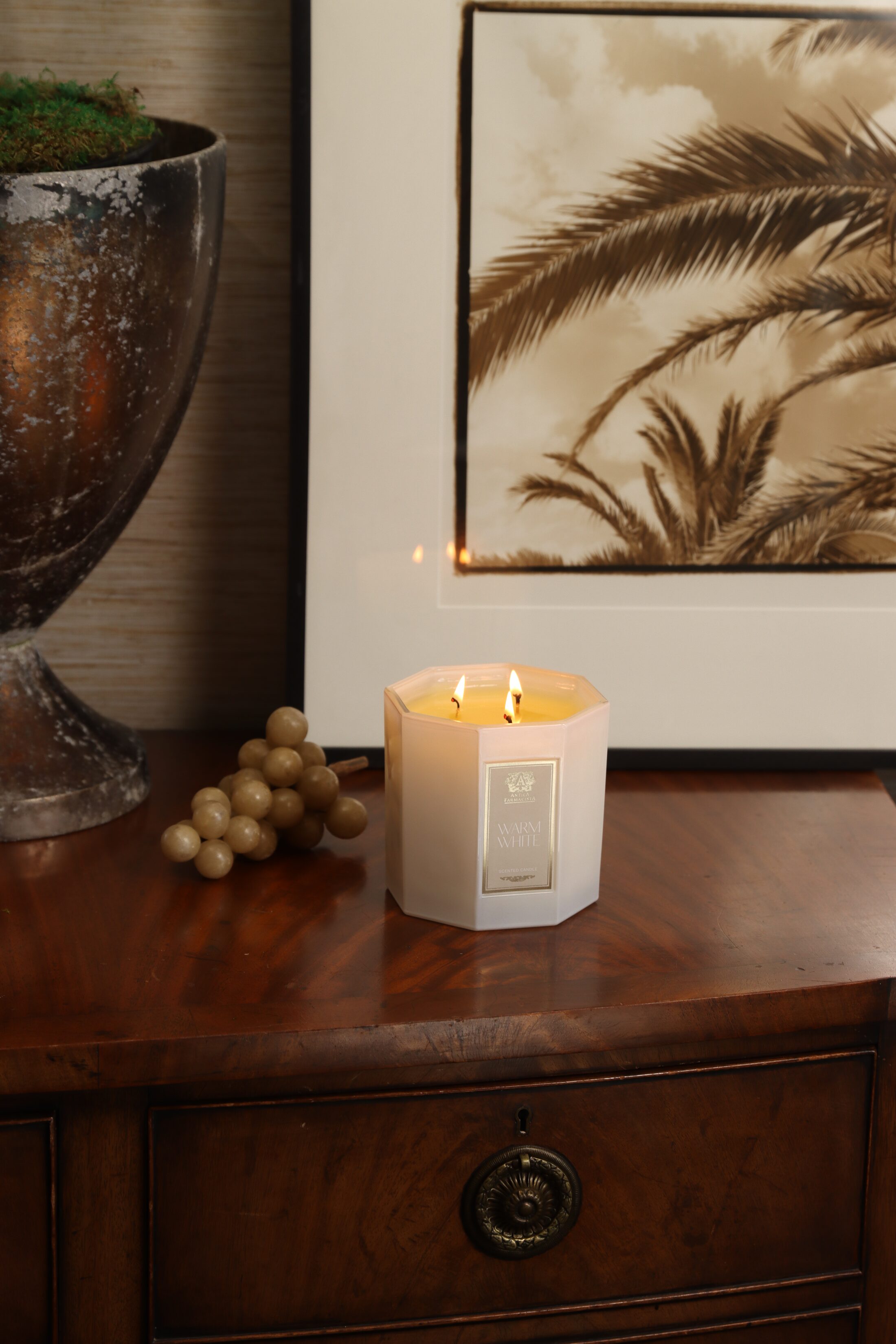 The Warm White candle lit on an antique mahogany chest beneath a sepia palm print, a sprig of champagne grapes beside it.