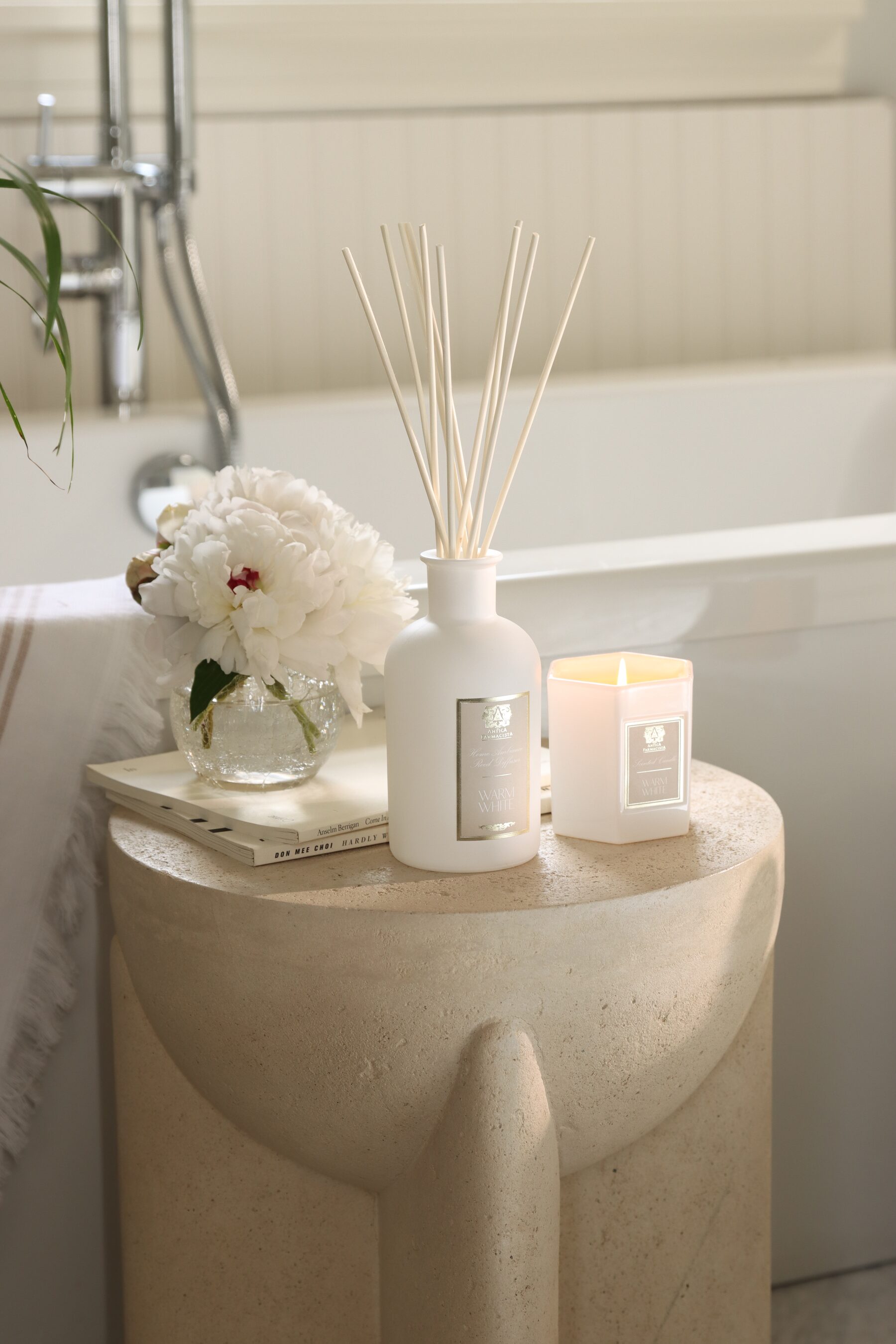 The opaque white Warm White diffuser and candle on a sculptural travertine stool at the bath’s edge, peonies beside them.