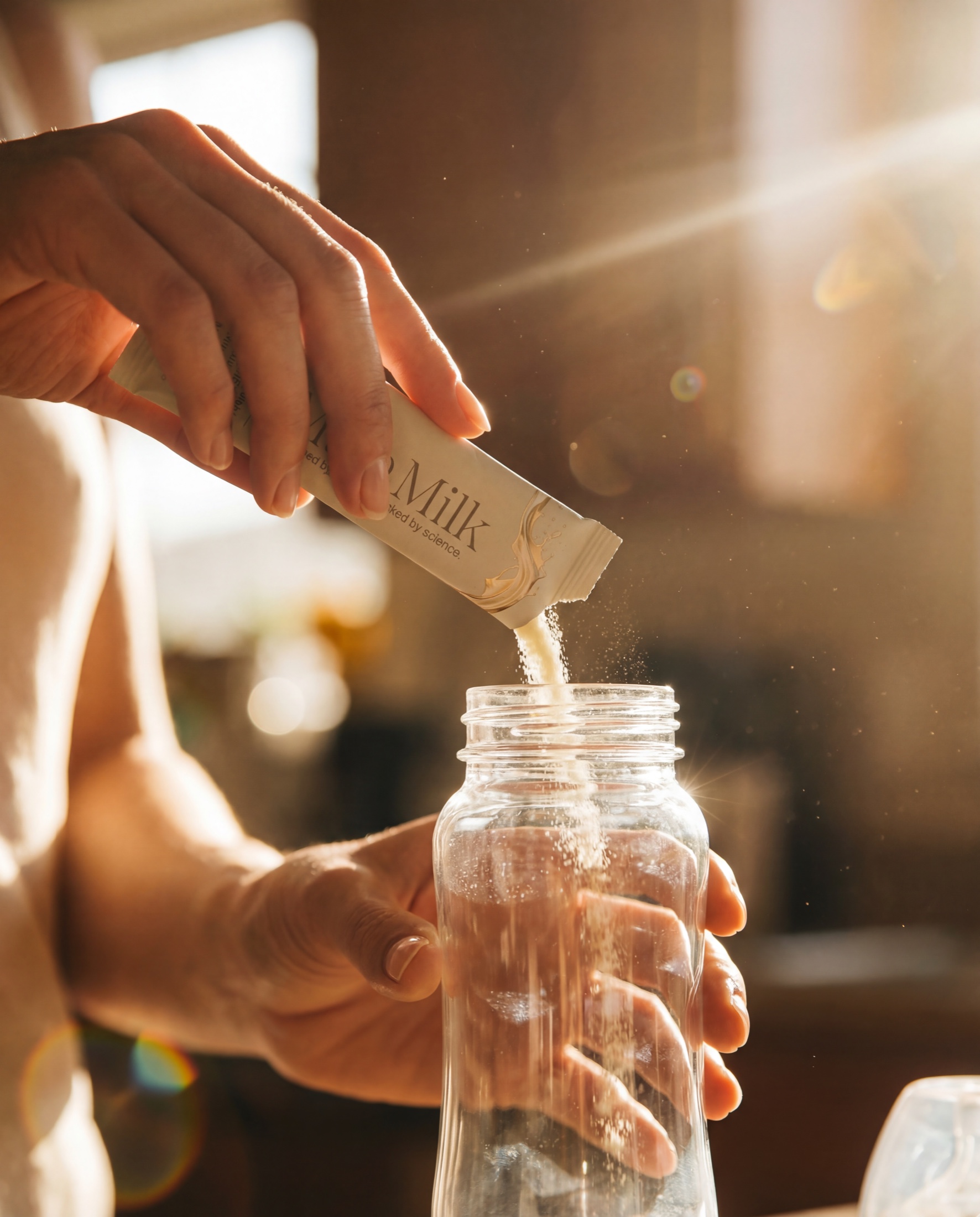 A hand pours MaiaMilk powder from a packet into a glass jar in warm backlight.